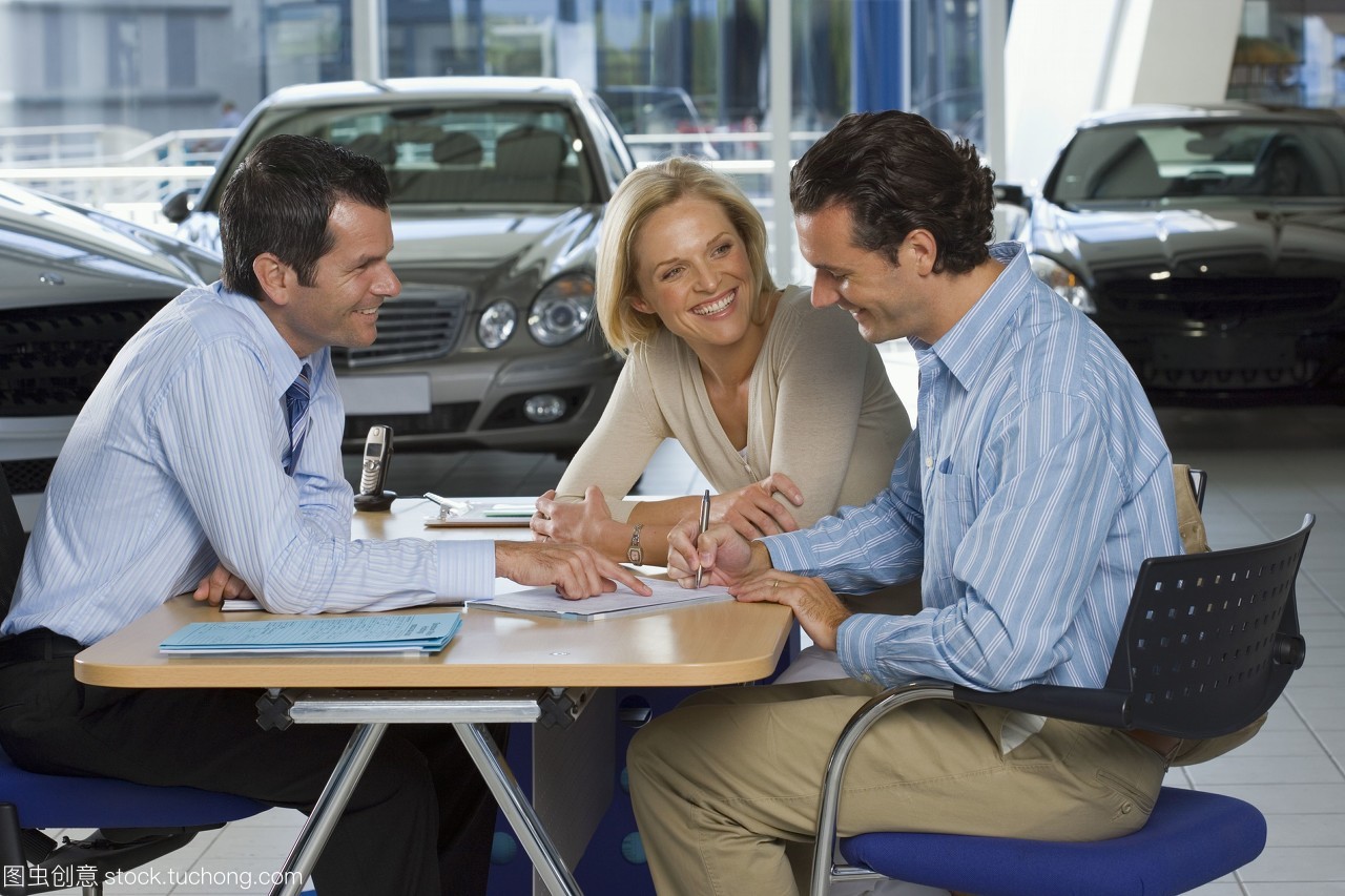 汽车销售员和夫妇坐在大汽车展示厅的办公桌前,签字文书,微笑,侧视